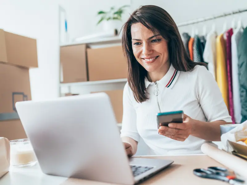Woman smiling while looking at a laptop and holding a cell phone in her left hand.